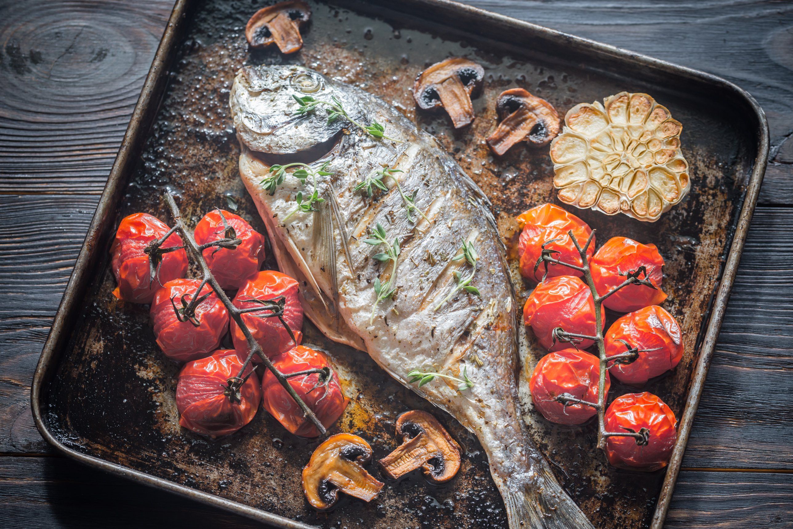 A whole sea bream baked on a tray with vine tomatoes, a halved head of garlic, mushrooms and thyme