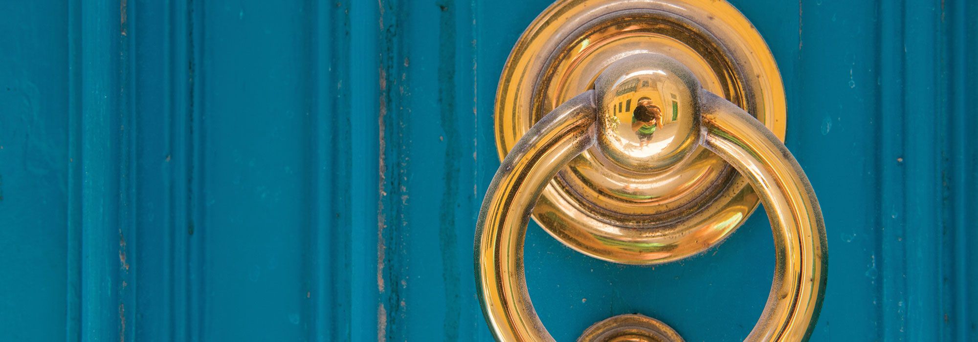 A polished brass ring knocker on a painted teal front door, the kind found on village houses across the Maltese islands