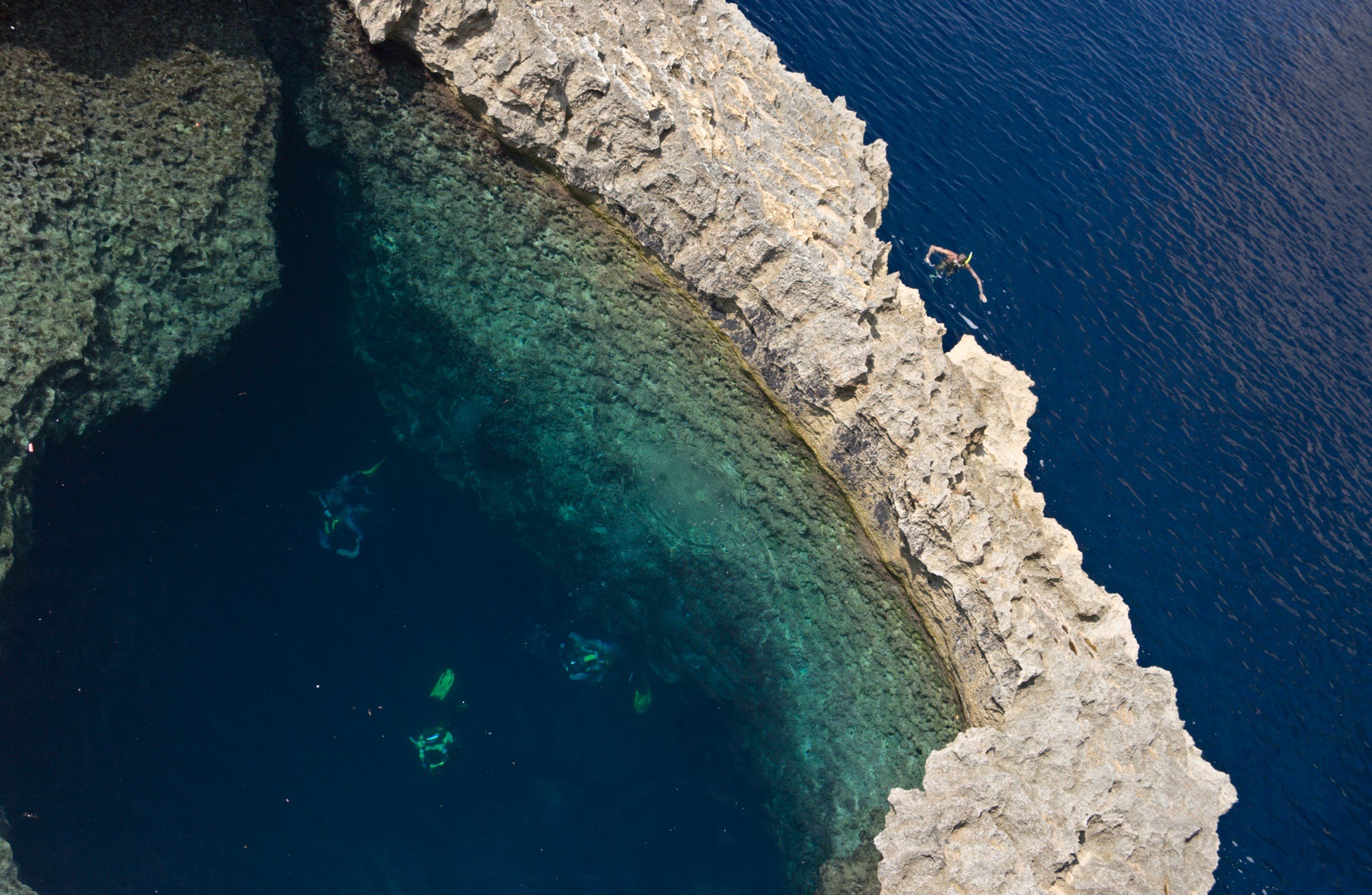 Looking straight down into the Blue Hole at Dwejra, with divers in the clear water and a snorkeller on the open sea beyond the rock