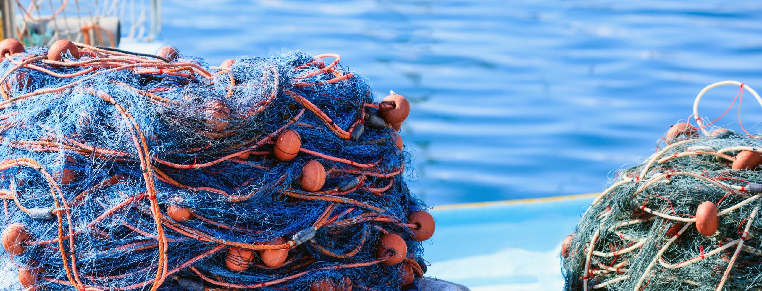 Bundled blue fishing nets with orange floats and rope stacked on a quayside beside clear water
