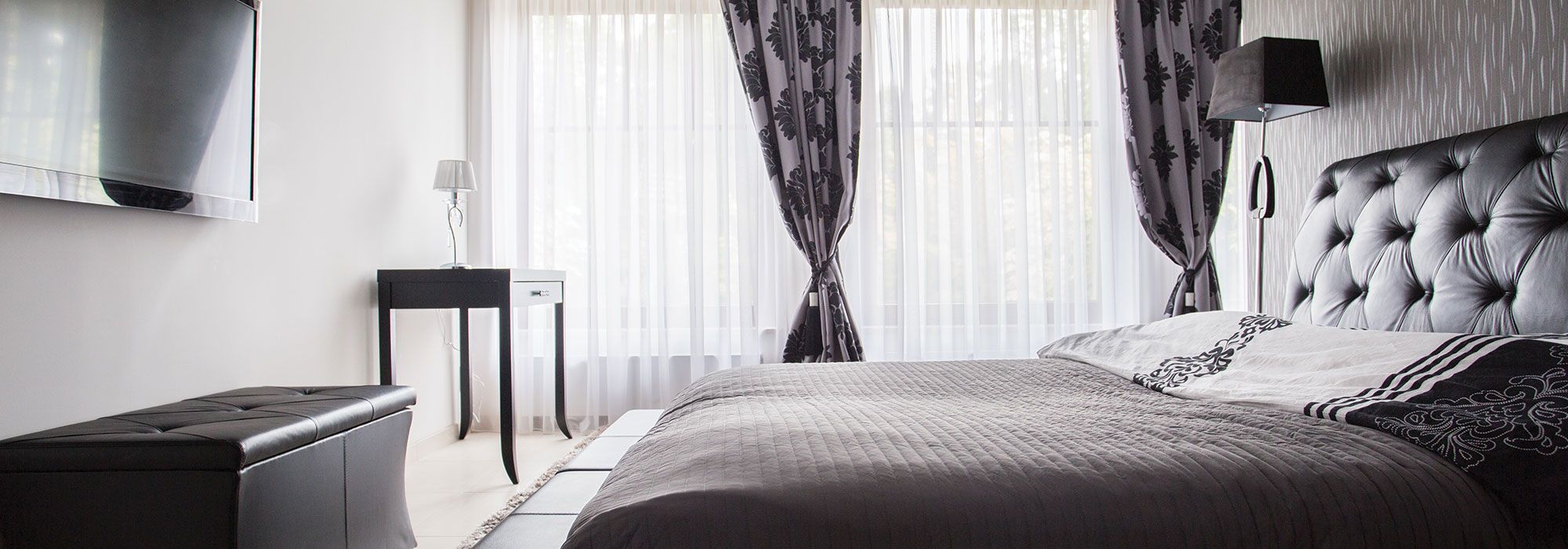 A bedroom with white walls, long sheer curtains and a dark upholstered bed, of the kind let as self-catering accommodation